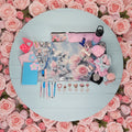 A styled flat-lay of various pink and floral-themed stationery and accessories arranged on a light gray surface against a white wall. The collection includes a floral print desk mat, pink notebooks, pens, heart-shaped clips, a pink water bottle, a zip pouch, and other cute stationery items.